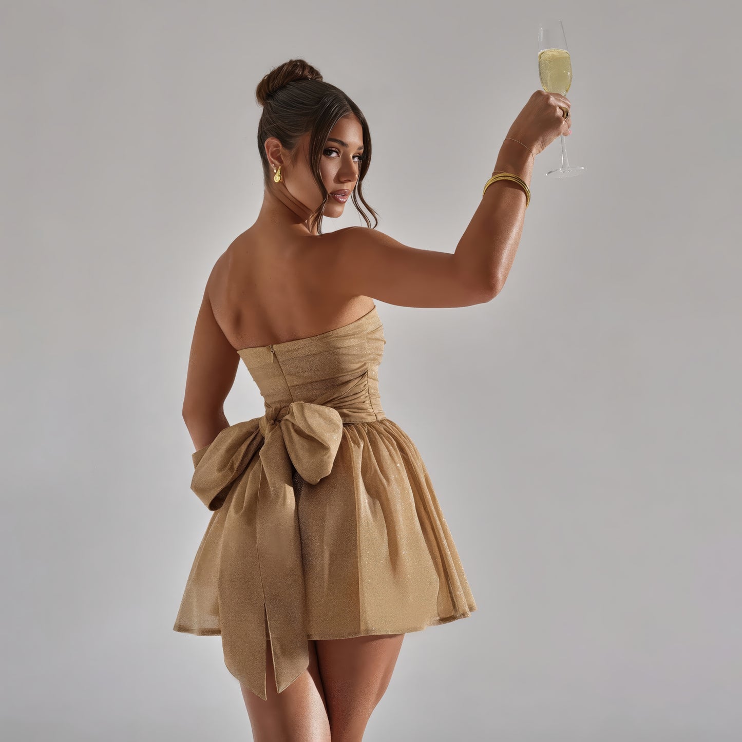 A woman in a strapless gold dress with a bow at the back is holding a glass of champagne and smiling.