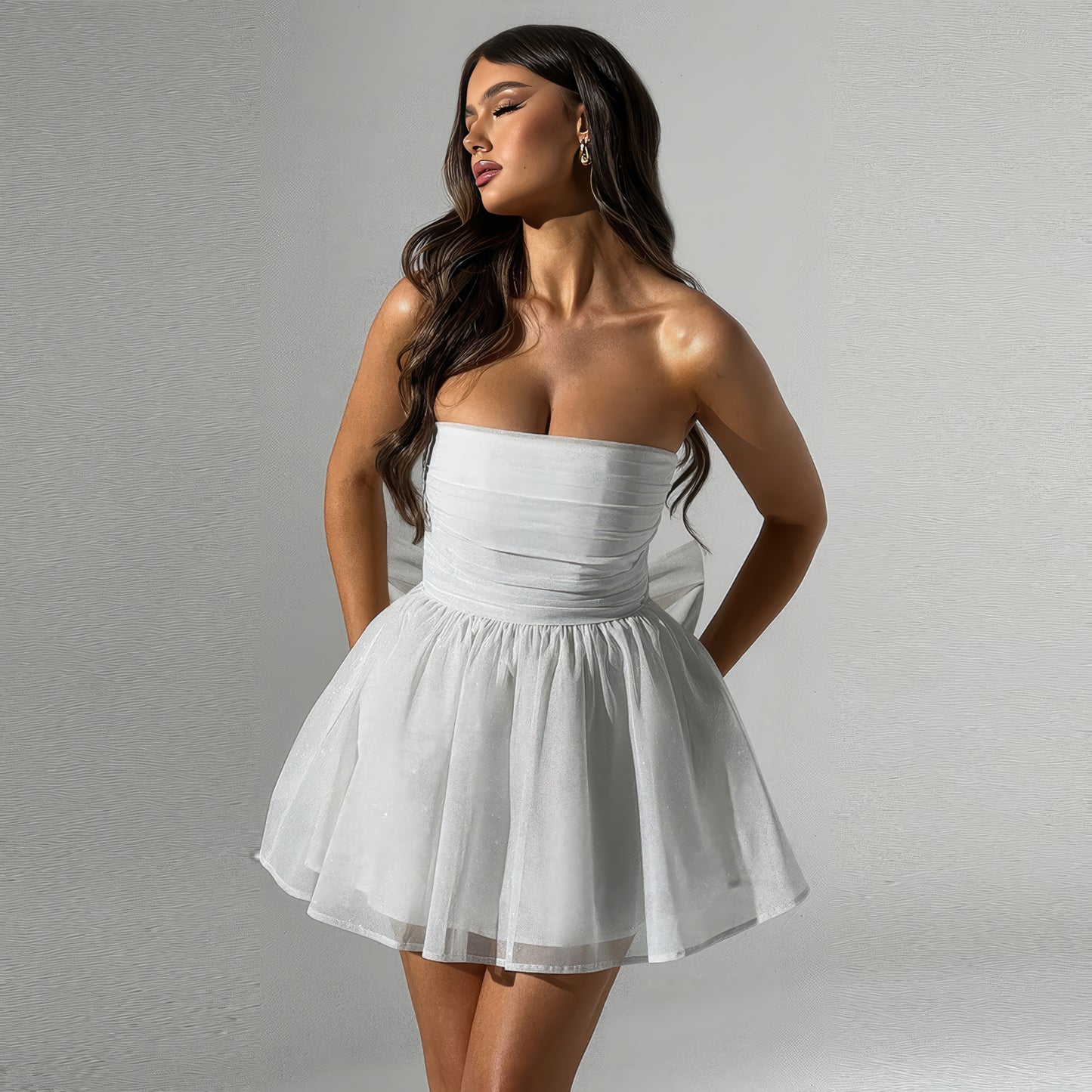 A woman wearing a strapless white dress with a full skirt, posing with her hands in her pockets against a plain background.